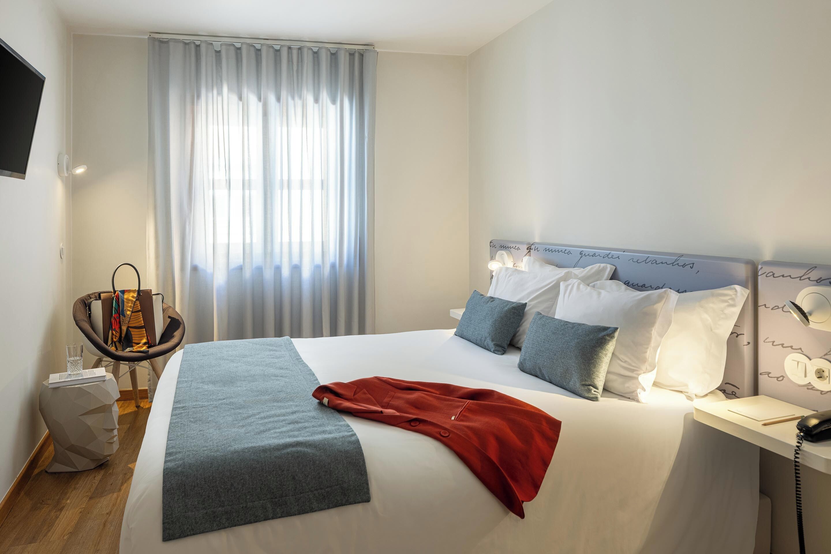 Habitació d'hotel amb un llit, una taula de nit amb una làmpada, una cadira, una televisió i una finestra amb cortines.