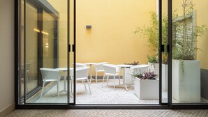Puerta de cristal que da a un patio con sillas y mesas blancas, una pared amarilla y plantas en macetas.