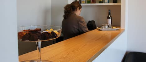 Una zona de bar con un bol de postre de vidrio transparente sobre una encimera de madera, una mujer sentada en la barra y estantes repletos de diversas botellas de licor al fondo.