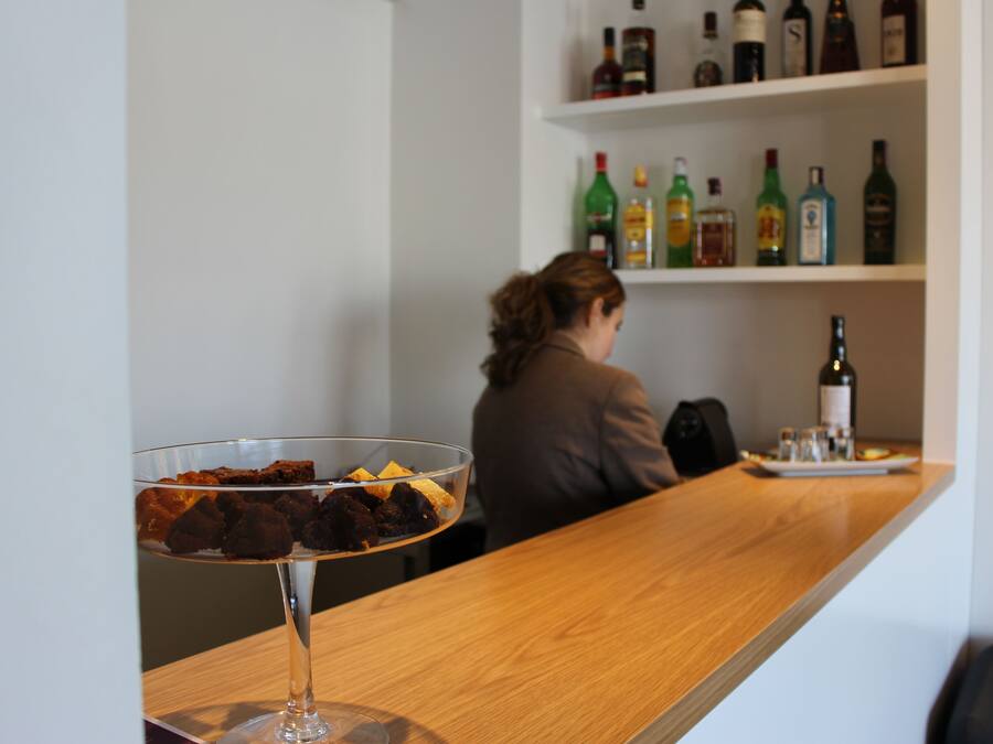 Uma área de bar com uma taça de sobremesa de vidro transparente sobre um balcão de madeira, uma mulher sentada no balcão, e prateleiras repletas de garrafas de bebidas alcoólicas ao fundo.