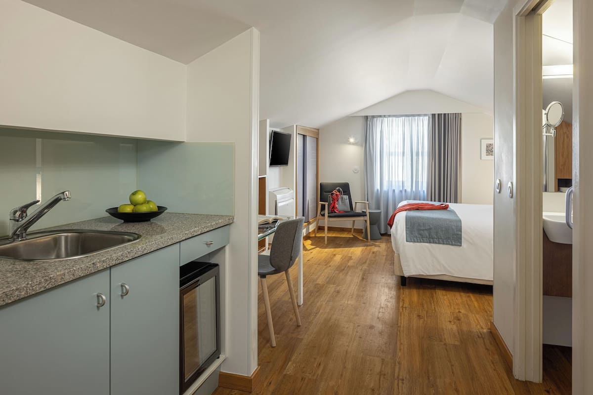 A compact hotel room with a kitchenette, a bed, a desk, and a bathroom.