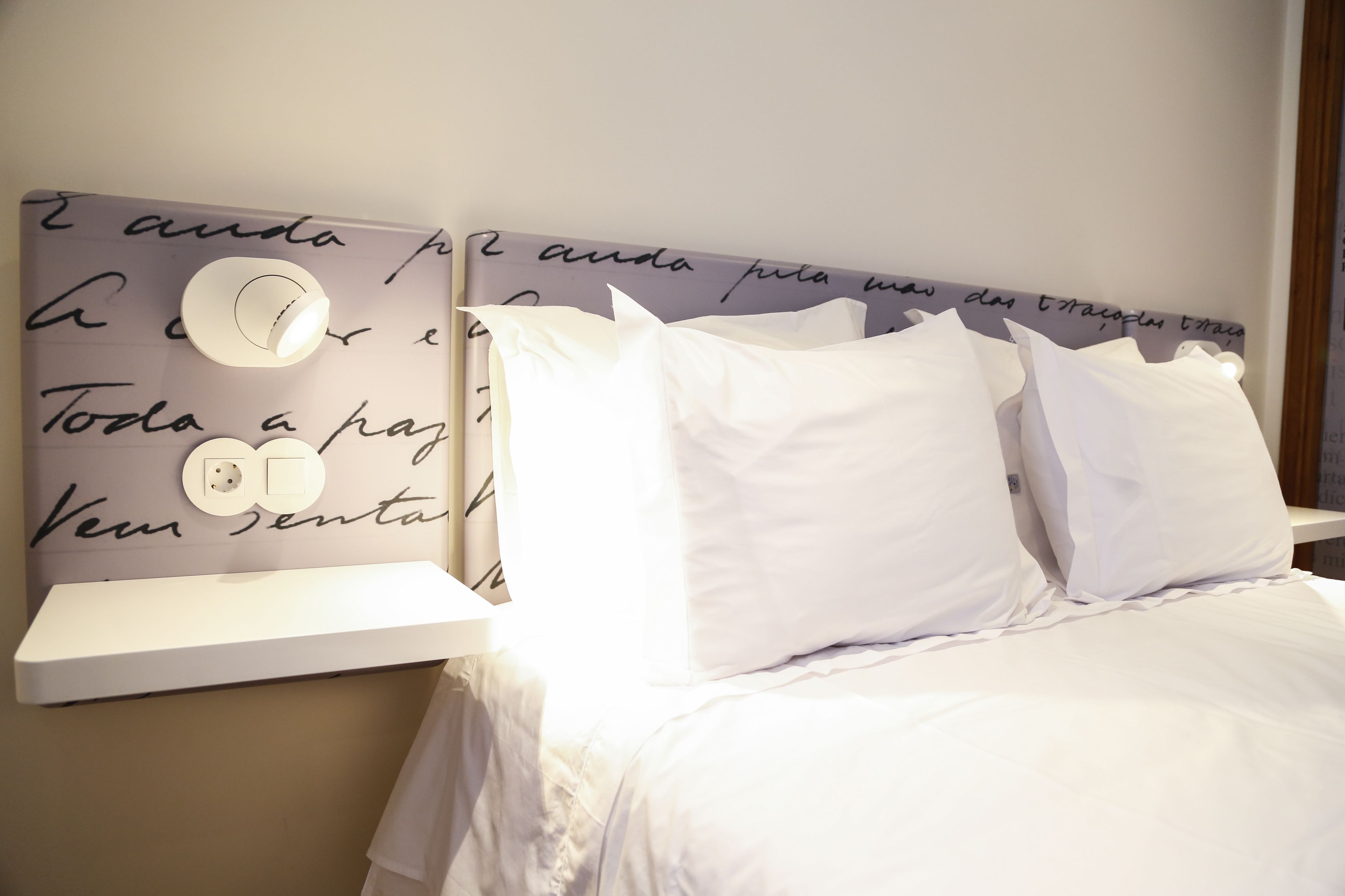 Una cama bien hecha con ropa de cama y almohadas blancas, un cabecero con detalles decorativos y una mesita de noche con una lámpara.