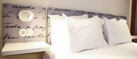 Una cama bien hecha con ropa de cama y almohadas blancas, un cabecero con detalles decorativos y una mesita de noche con una lámpara.