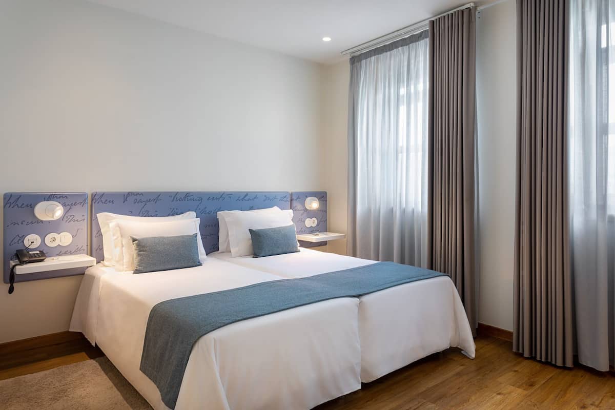 A neatly made double bed with a blue blanket, two bedside tables with lamps, a wall with decorative writing, and a window with curtains.