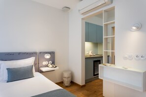 A modern hotel room with a bed, a small desk with a phone, a mirror, and a kitchenette.