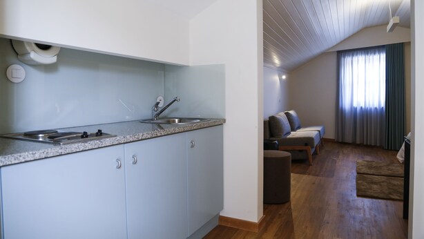 A modern kitchen with a built-in oven and sink, a dining area with a wooden table and chairs, and a living room with a sofa and a large window. - Hotel das Salinas (Aveiro)