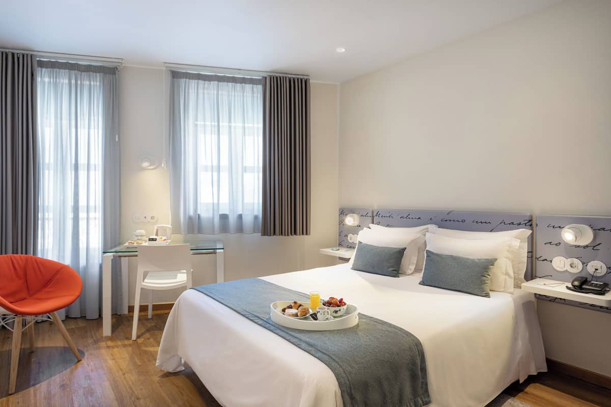 A hotel room with a bed, a desk, a chair, and a bedside table with a lamp.