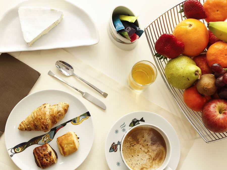 Desayuno con cruasanes, un sándwich, una cesta de fruta y una taza de café sobre una mesa.