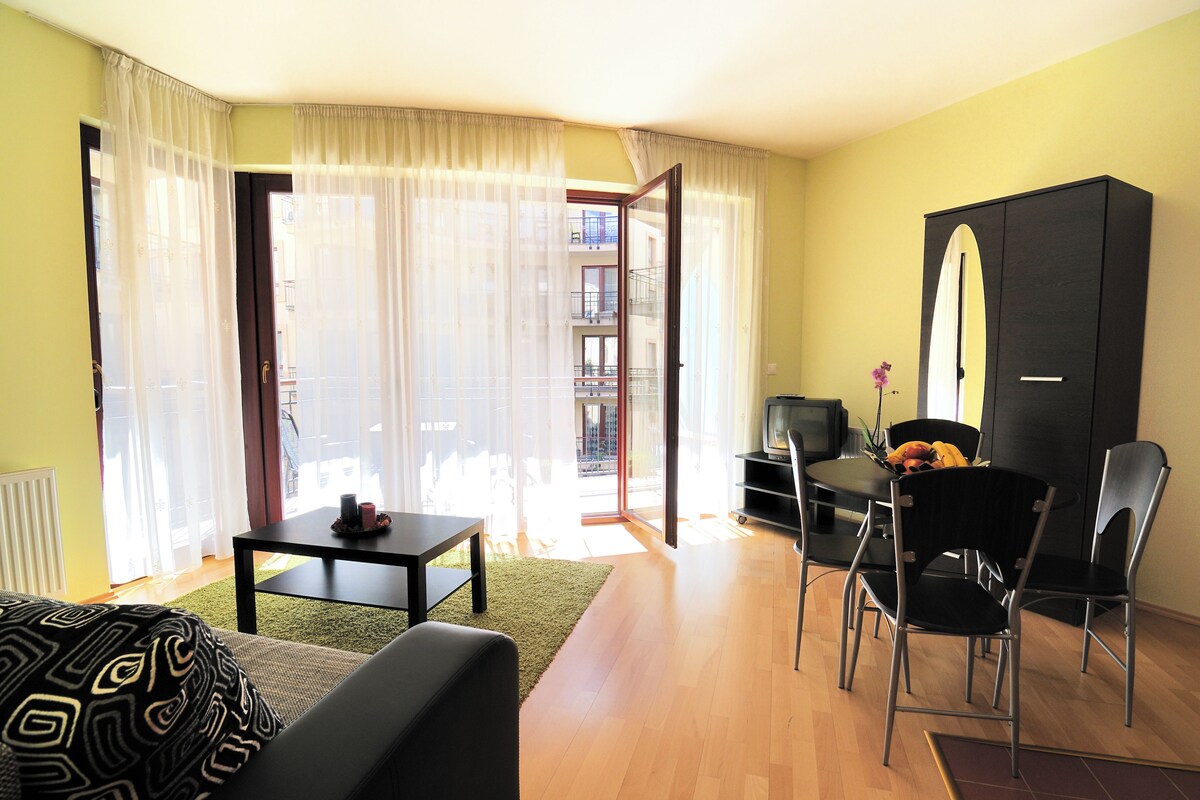 Una sala de estar moderna con un sofá, una mesa de comedor y sillas. Hay un televisor sobre un mueble bajo, un aparador y un balcón visible entre las cortinas.