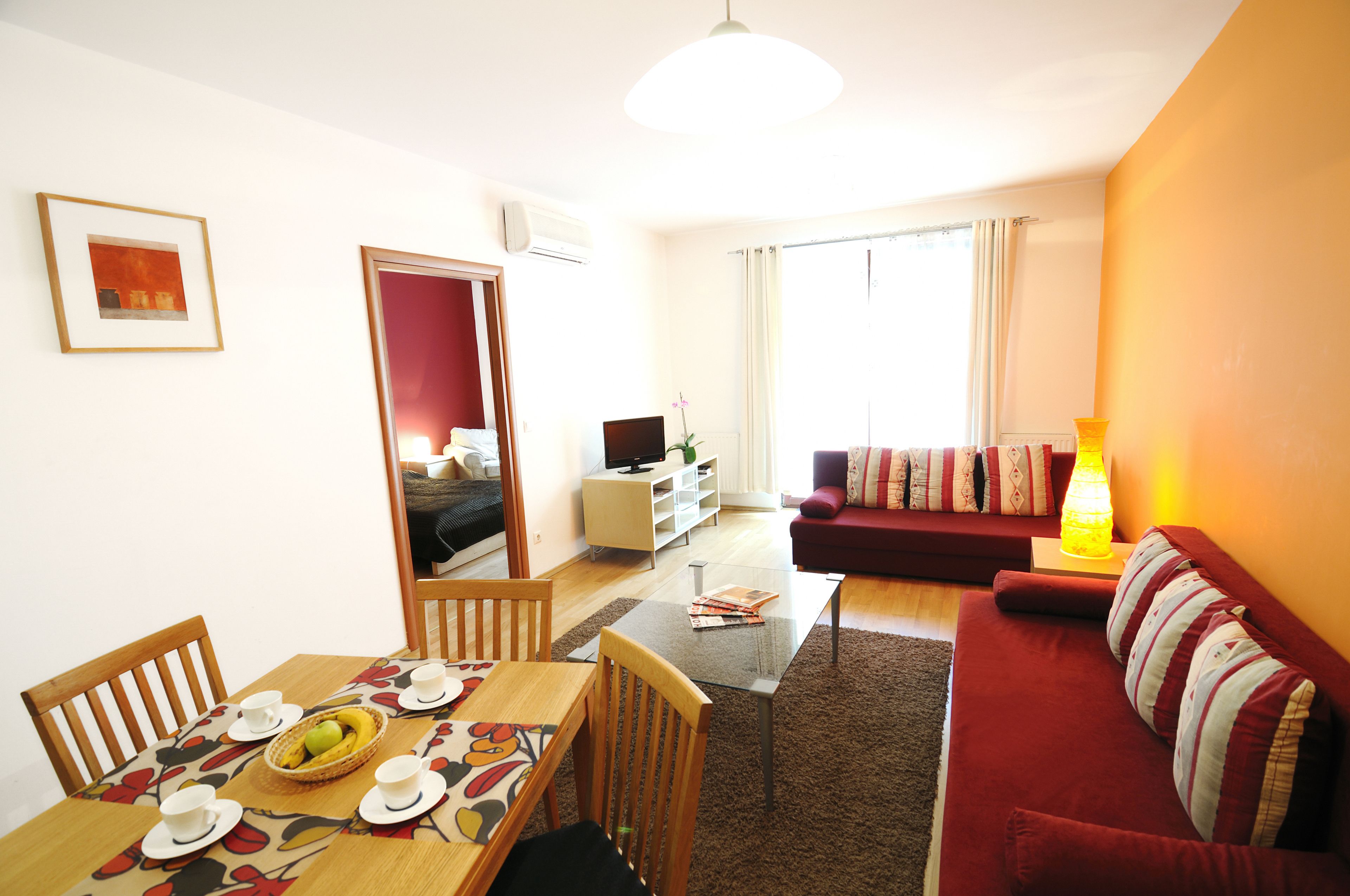 A cozy living room with a red sofa, a dining table with chairs, and a TV in the background.