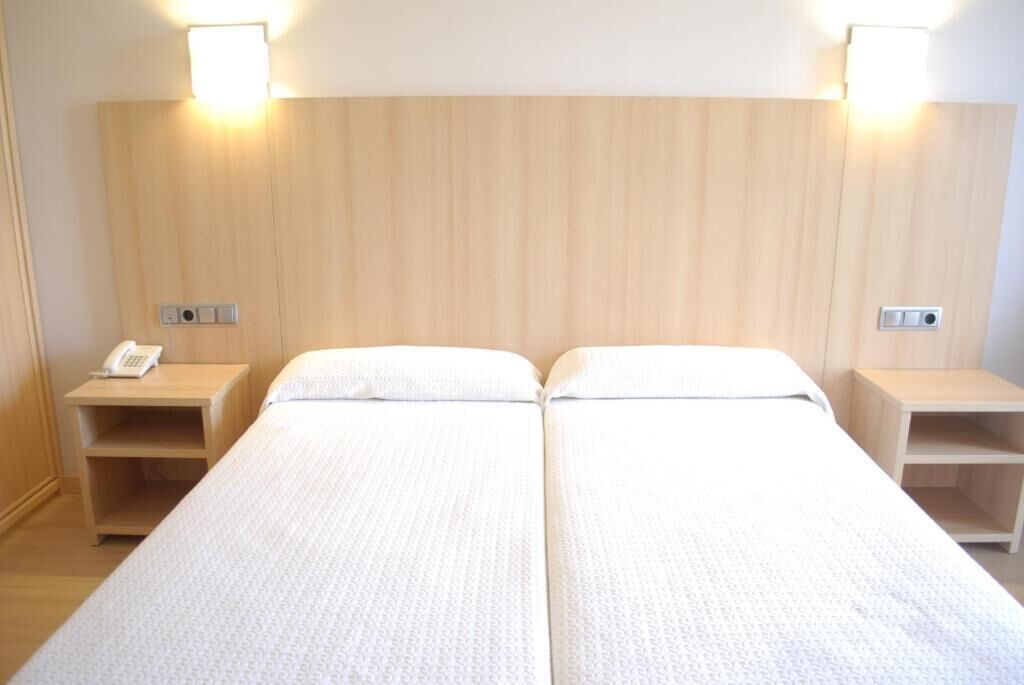 Habitación de hotel con dos camas individuales, cabecero de madera, mesitas de noche con lámparas y un teléfono en una de las mesitas.