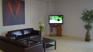 Un salón con un sofá de cuero oscuro, una mesa de centro de vidrio, un televisor sobre un mueble de madera y una planta en maceta.