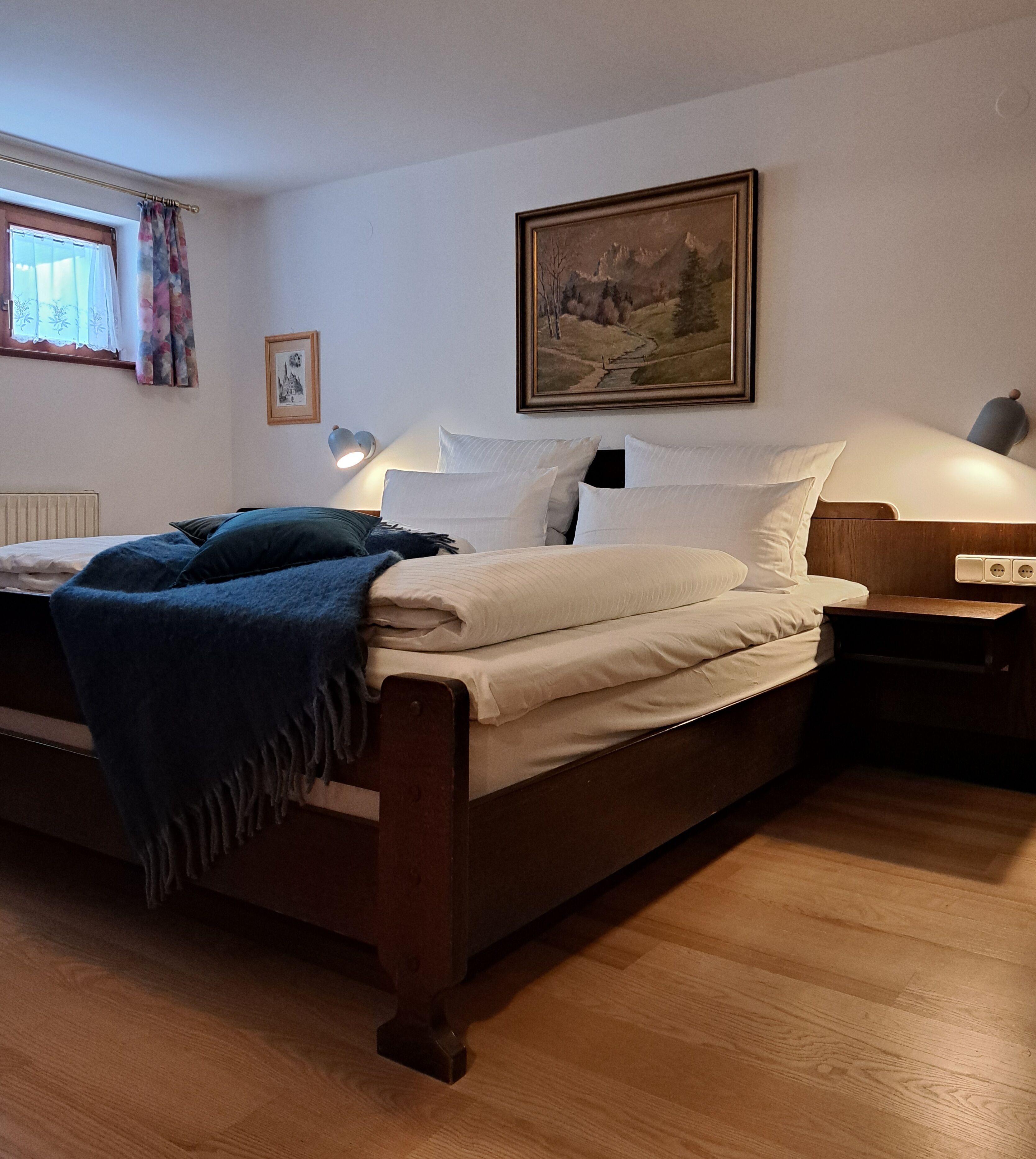 Ein Schlafzimmer mit einem hölzernen Bett, weißer Bettwäsche, einem Bild an der Wand, einem Nachttisch mit Lampe und einem Fenster mit Vorhängen.