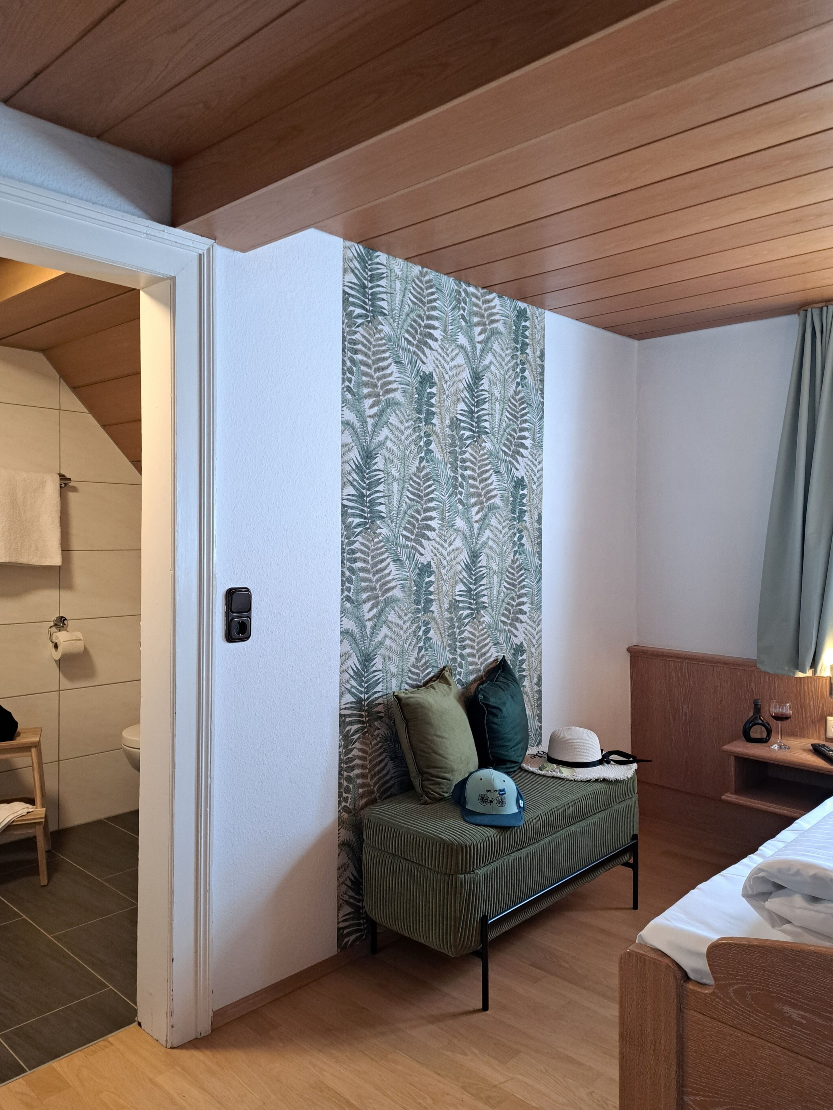 Habitación de hotel con techo de madera, un sofá verde con almohadas, una mesita con una botella y un vaso, y un baño visible a través de una puerta abierta.