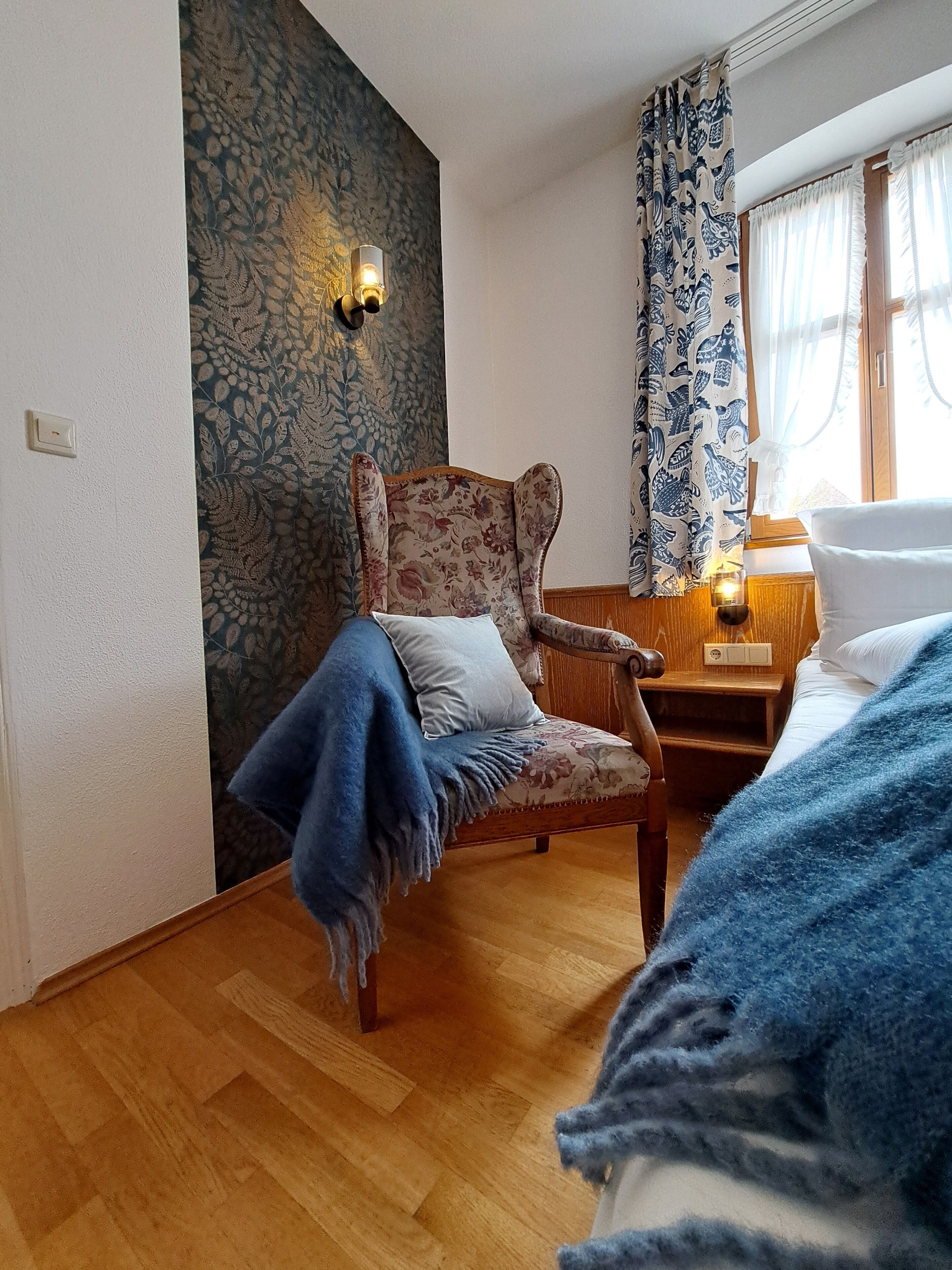Ein Zimmer mit Holzboden, einem blumigen Sessel, einem Bett mit weißer Bettwäsche und einer Wand mit blattbedecktem Wandpapier.
