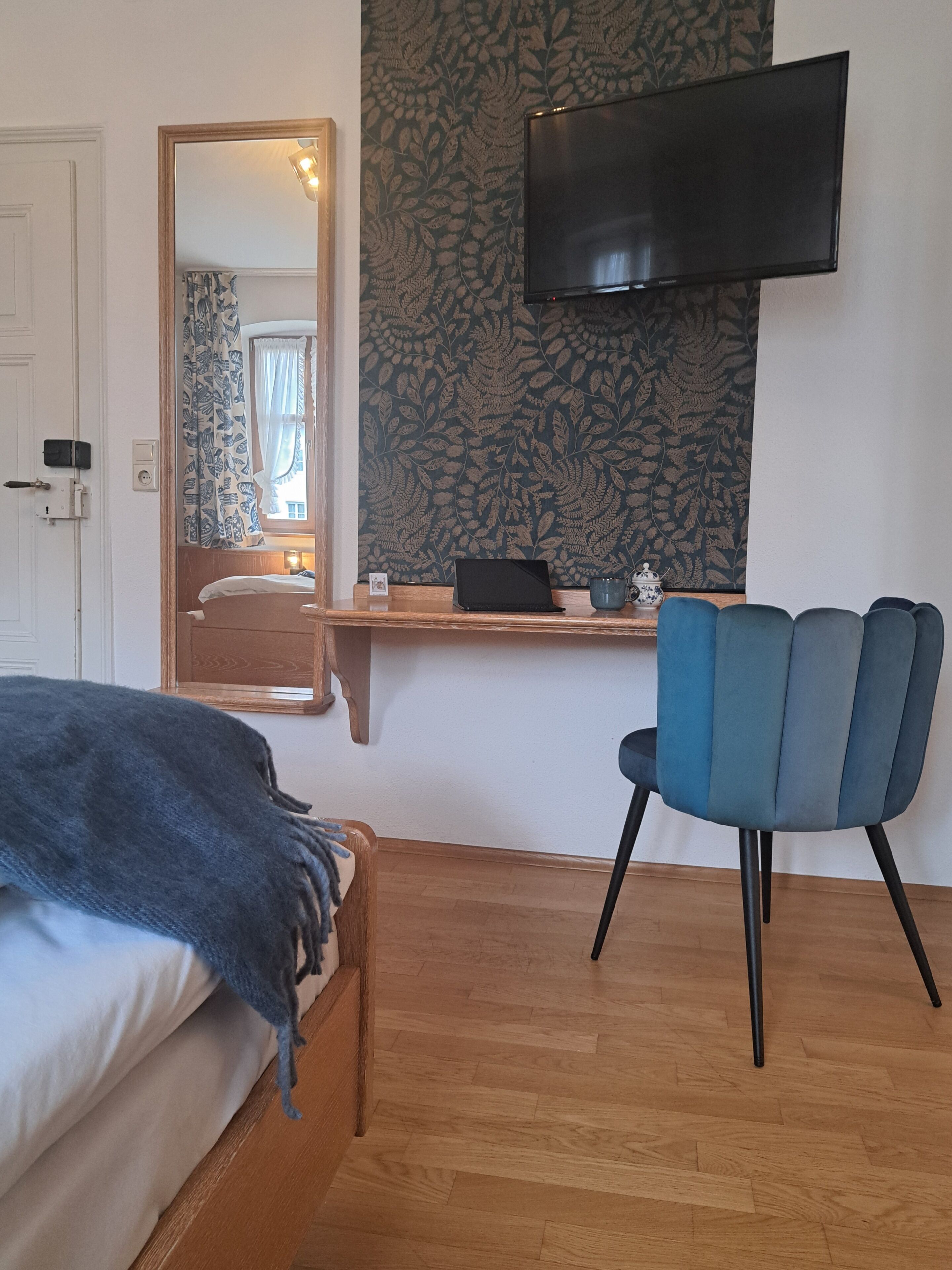 A bedroom with a bed, a desk with a laptop, a chair, a TV on the wall, and a mirror.
