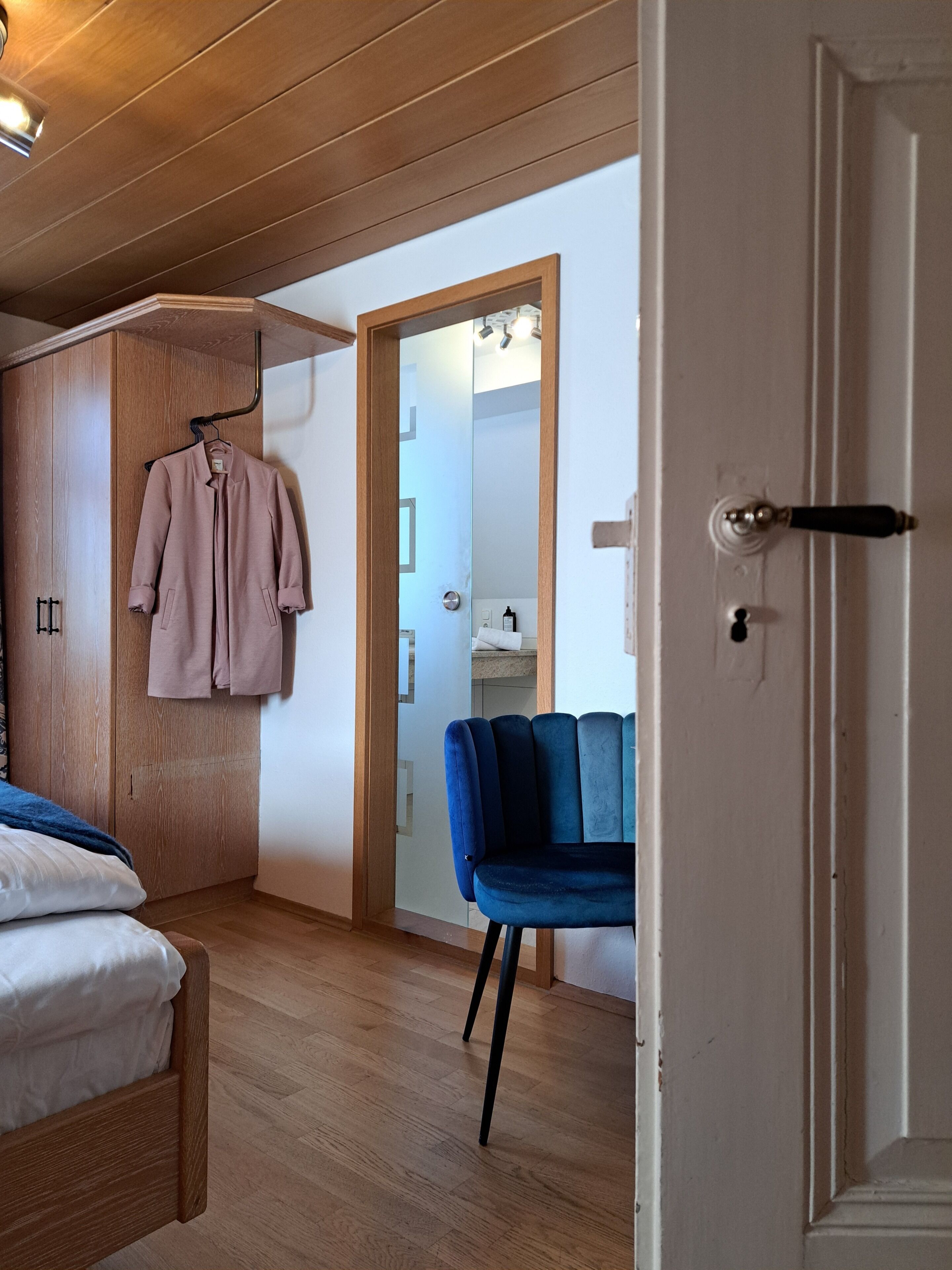 A bedroom with a bed, a wardrobe, a hanging coat, and a bathroom visible through a door.