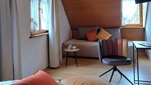 A cozy room with a wooden ceiling, a sofa with cushions, a chair, a small table, and a window with curtains. - Gasthof Hotel Bezold (Rothenburg ob der Tauber)