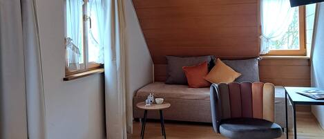 Una habitación acogedora con techo de madera, un sofá con cojines, una silla, una mesita y una ventana con cortinas.