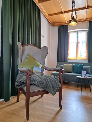Ein Holz-Sessel mit Kissen, ein kleiner Tisch mit einer Speisekarte und eine Couch mit Kissen in einem Zimmer mit Holzboden und einem Fenster mit Vorhängen.