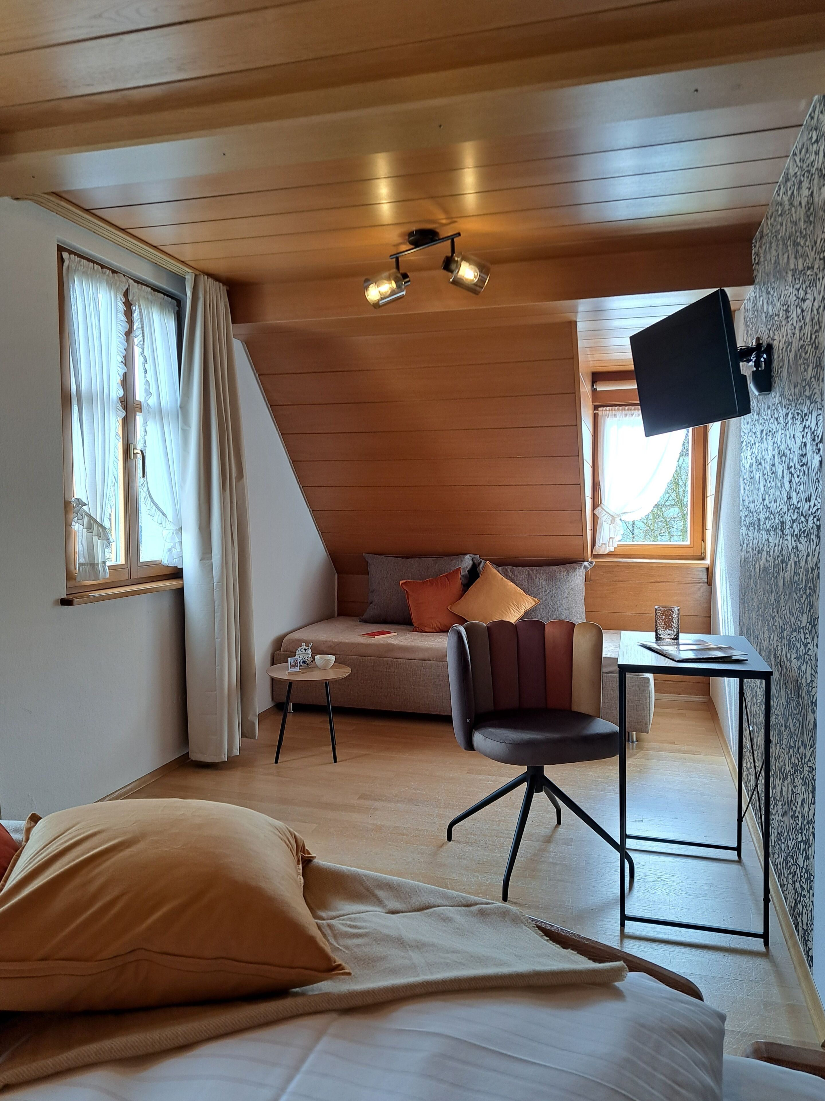 A cozy room with a bed, a small table, a chair, a sofa, a TV, and a window with curtains.