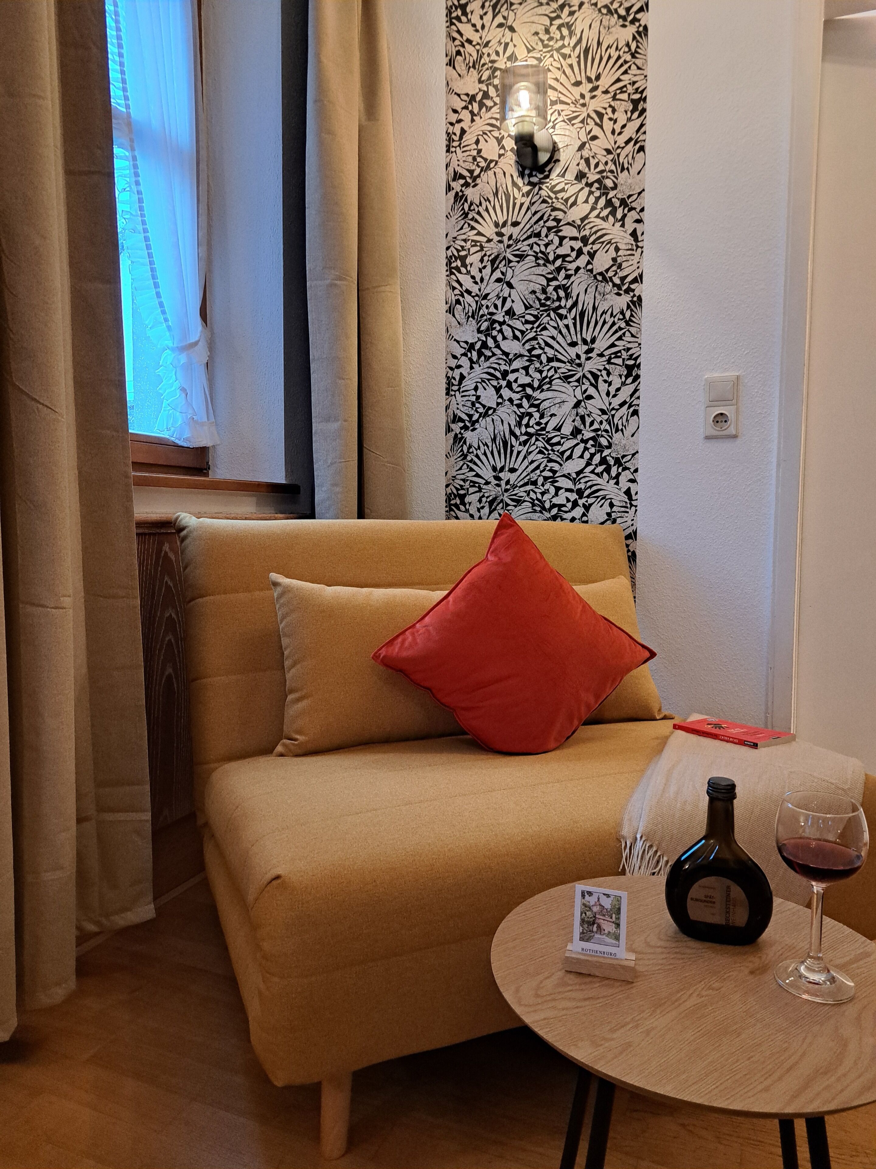 Una habitación acogedora con un sofá, una mesita redonda con una botella y un vaso, y una pared con estampado floral.