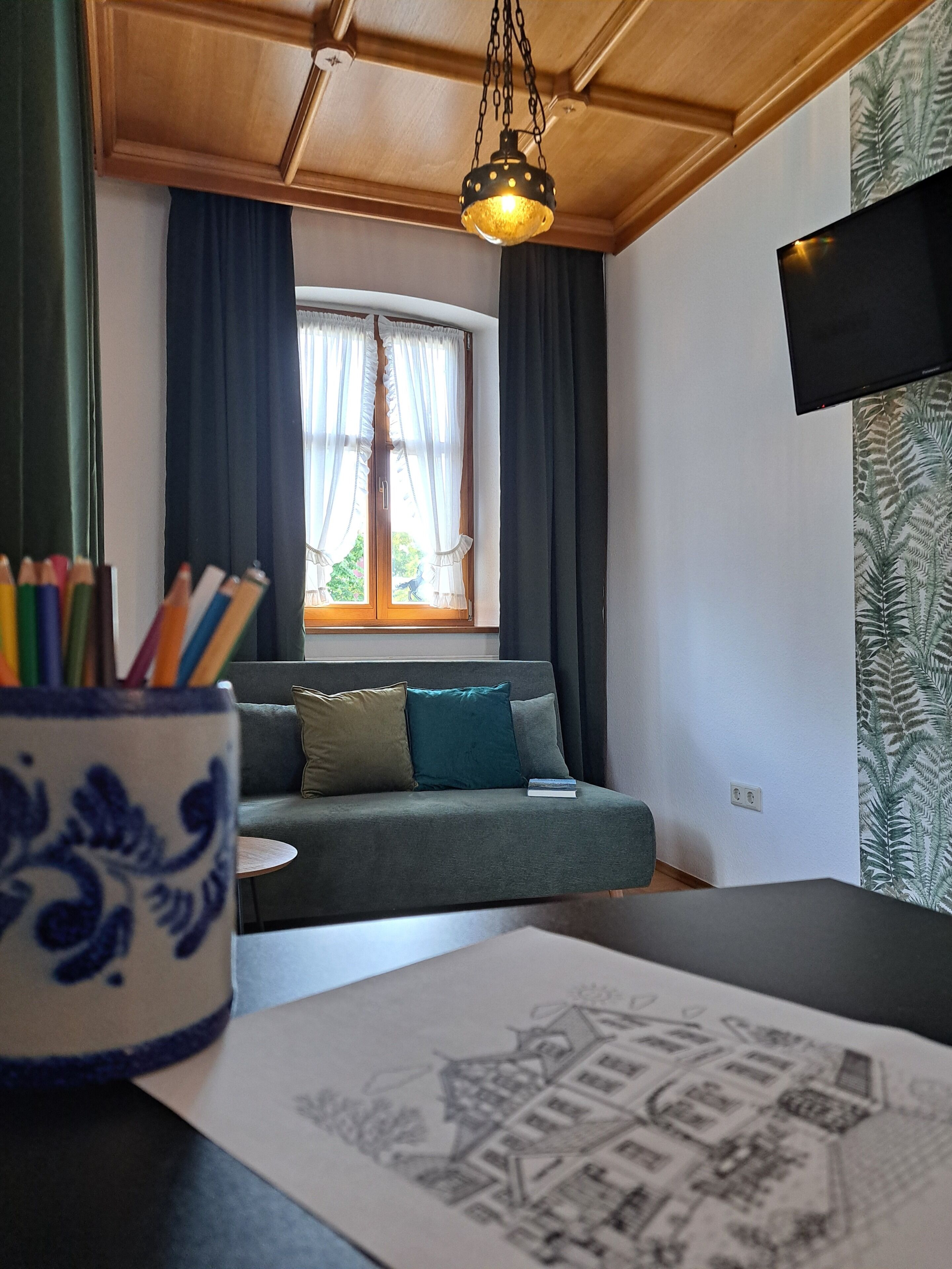 Una habitación con un sofá, una mesa con un dibujo, un jarrón blanco y azul con motivos, una lámpara colgante y una ventana con cortinas.