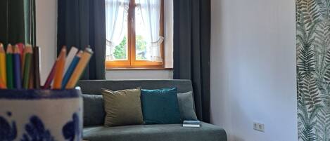 Una habitación con un sofá, una mesa con un dibujo, un jarrón blanco y azul con motivos, una lámpara colgante y una ventana con cortinas.
