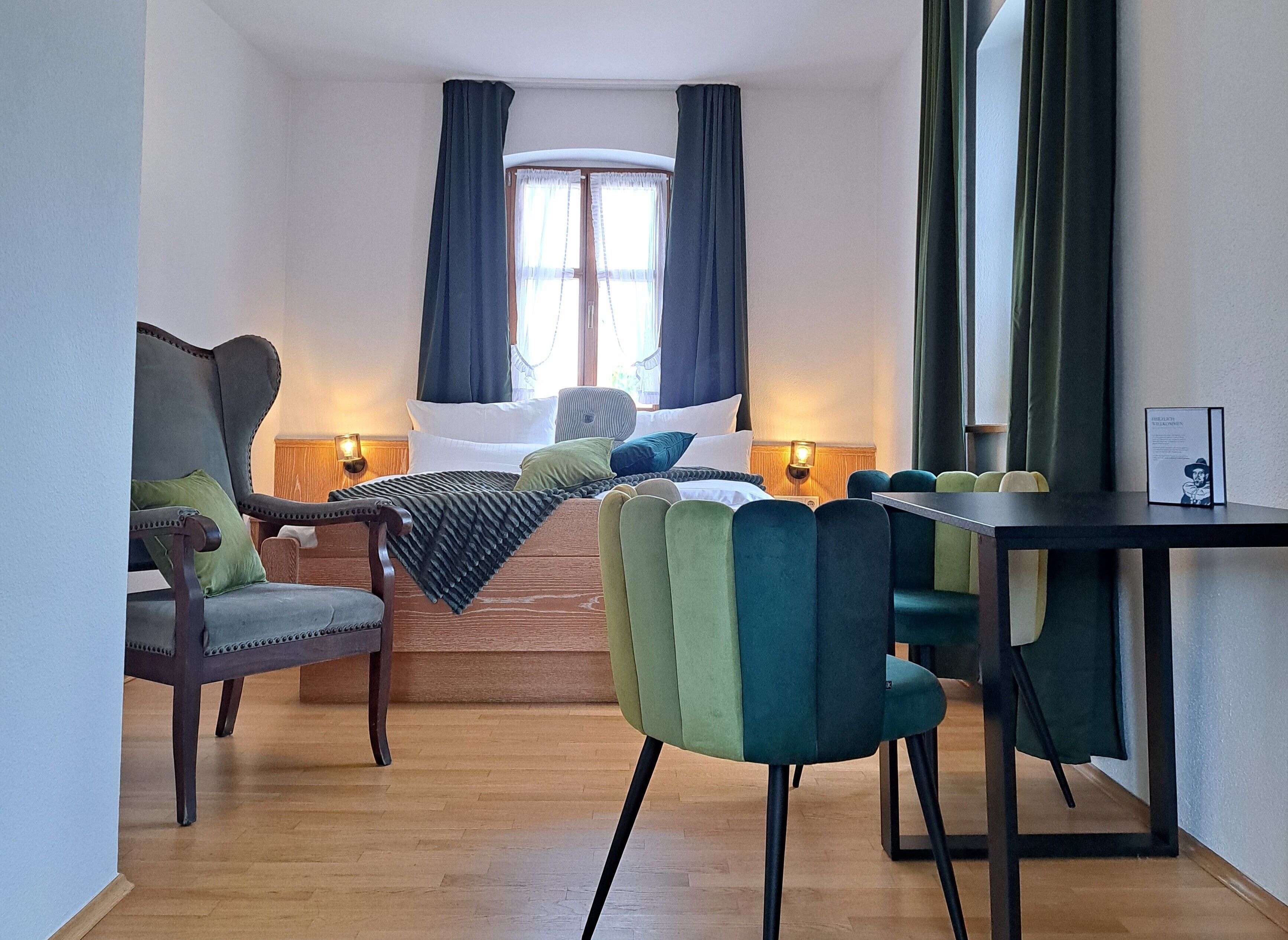 Habitación de hotel con cama, escritorio, dos sillas, ventana con cortinas y lámpara fijada a la pared.