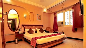 A hotel room with a large bed, a round mirror, a bedside table with a lamp, and a window with curtains. - Ratnawali (Jodhpur)