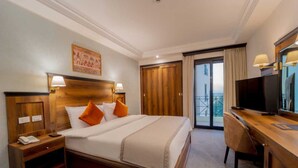A hotel room with a large bed, wooden headboard, two bedside tables with lamps, a desk with a chair, a television, and a balcony with curtains. - Hotel Morris Luxor (Luxor)