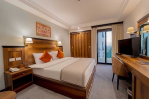 Habitación de hotel con una cama grande, cabecera de madera, dos mesitas de noche con lámparas, un escritorio con una silla, un televisor y un balcón con cortinas.