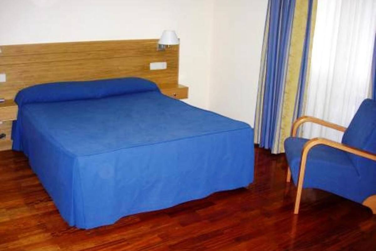 A room with a wooden headboard bed, a blue bedspread, a single bedside lamp, a blue chair, and a window with curtains.