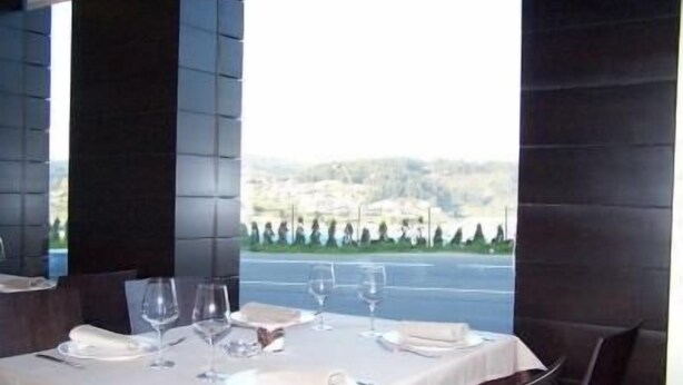 A dining table set with white tablecloth, plates, and wine glasses, with a view of a road and trees outside. - Hotel Arteixo (Arteixo)