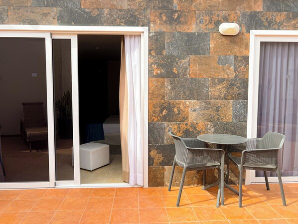 Una habitación con puerta de cristal corredera que da a un balcón, una mesita redonda con dos sillas y una planta en maceta.