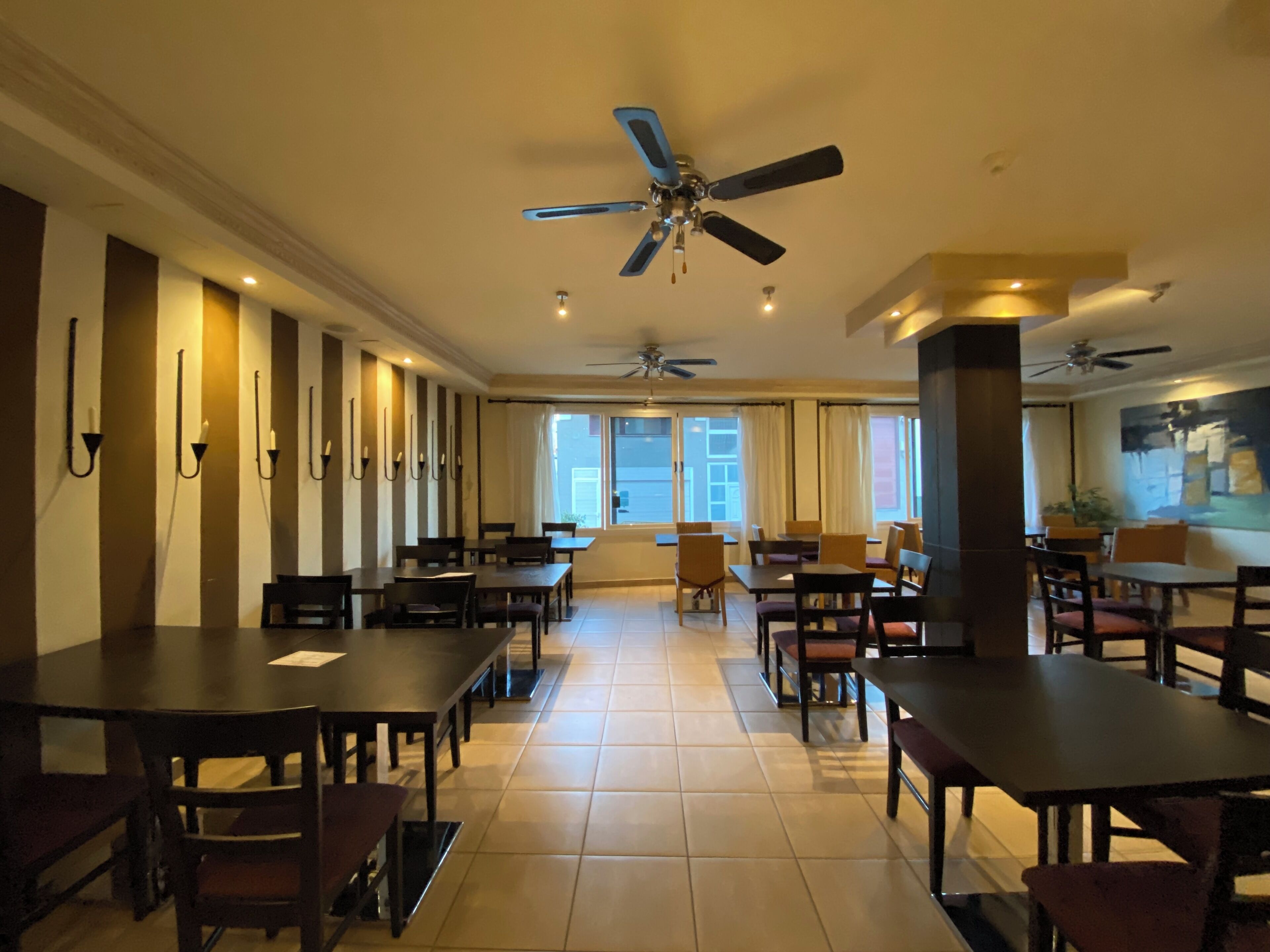 A spacious dining area with multiple tables and chairs, ceiling fans, and large windows allowing natural light.
