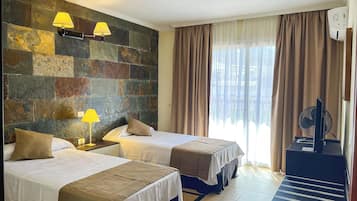 A hotel room with two beds, a stone wall, a window with curtains, and a standing fan.