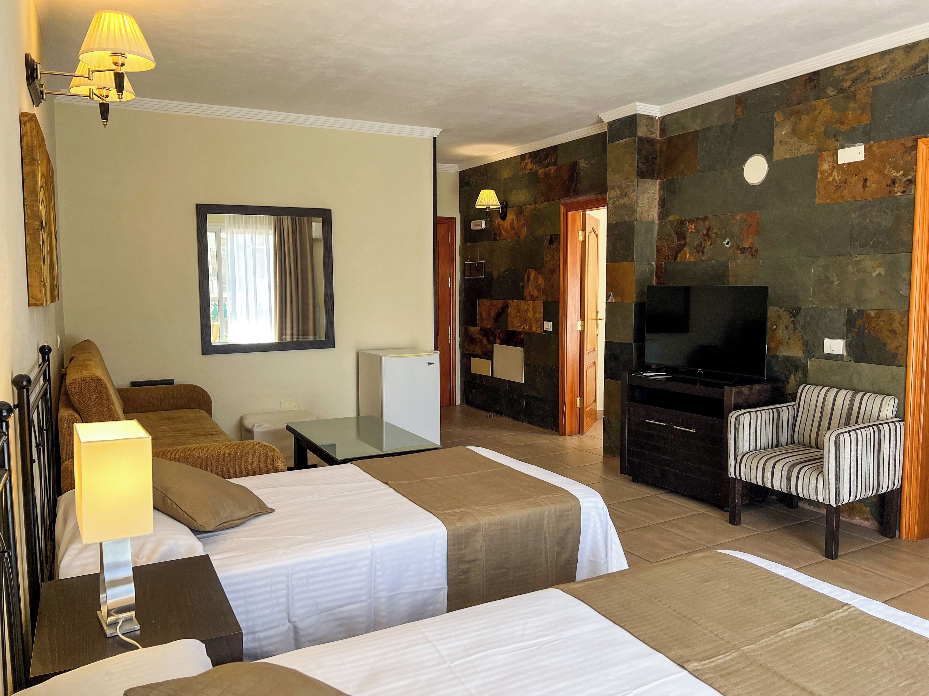 A hotel room with two beds, a television, a seating area with a striped chair, and a stone wall.