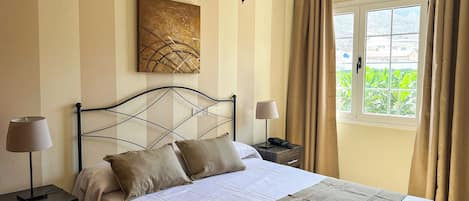 A neatly made bed with a headboard, two bedside lamps, a nightstand with a phone, a painting on the wall, and a window with curtains.