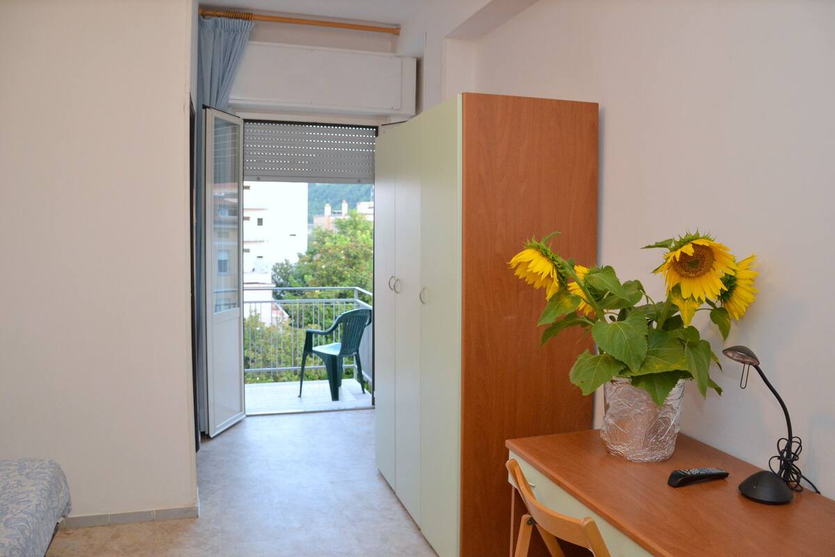A room with a wooden desk, a vase with sunflowers, a remote control, and a view of a balcony with a chair.