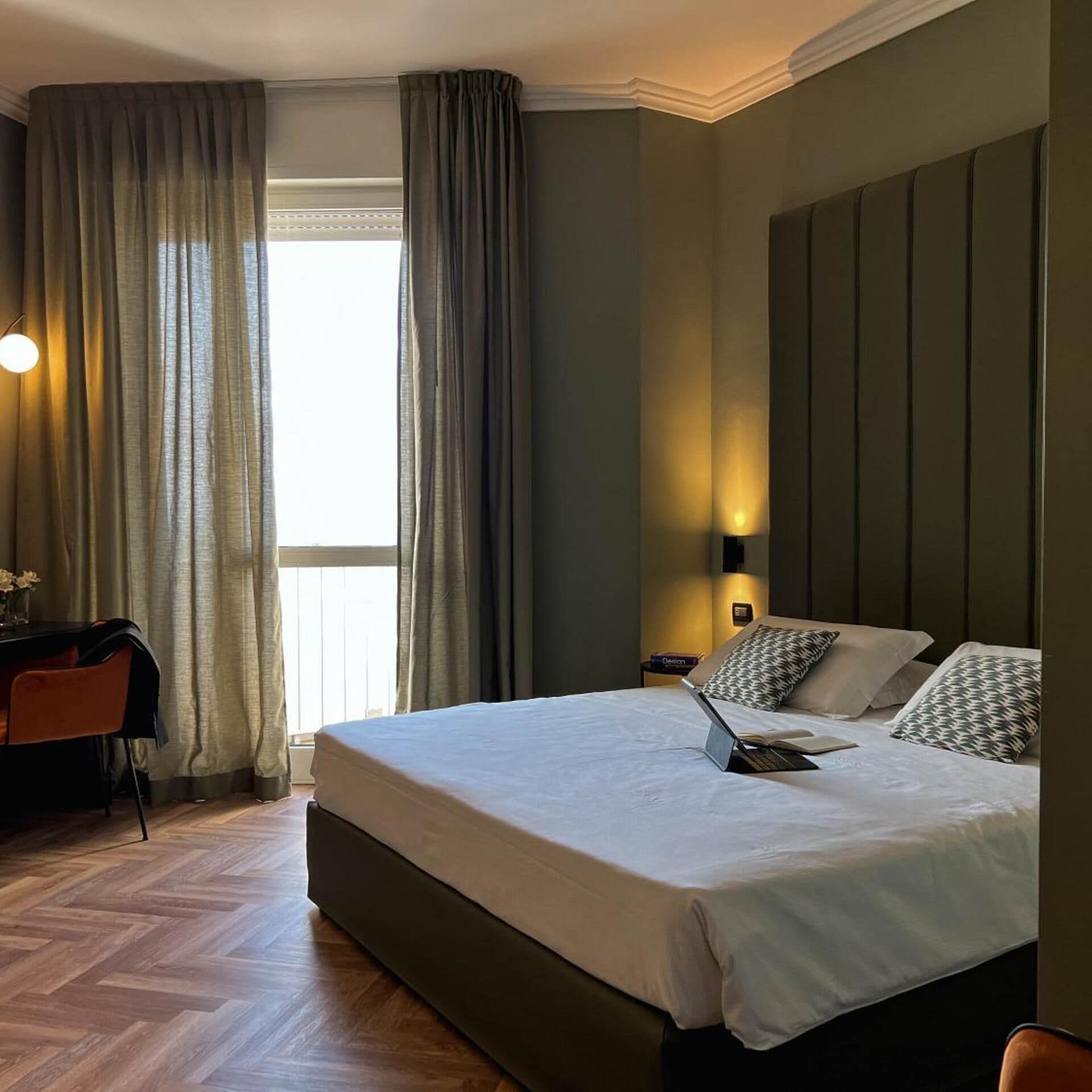 A hotel room with a large bed, a desk, and a chair. There is a window with curtains, a wall-mounted lamp, and a view of the outside.