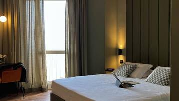 A hotel room with a large bed, a desk, and a chair. There is a window with curtains, a wall-mounted lamp, and a view of the outside.