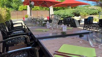 Een buitenterras met tafels gedekt voor een maaltijd, omgeven door rode parasols, zwarte stoelen en begroeiing op de achtergrond.