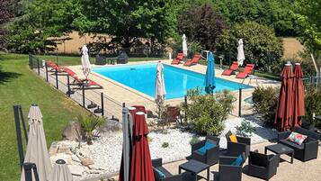 A rectangular swimming pool with red lounge chairs and umbrellas, surrounded by a fence, with a grassy area and trees in the background.
