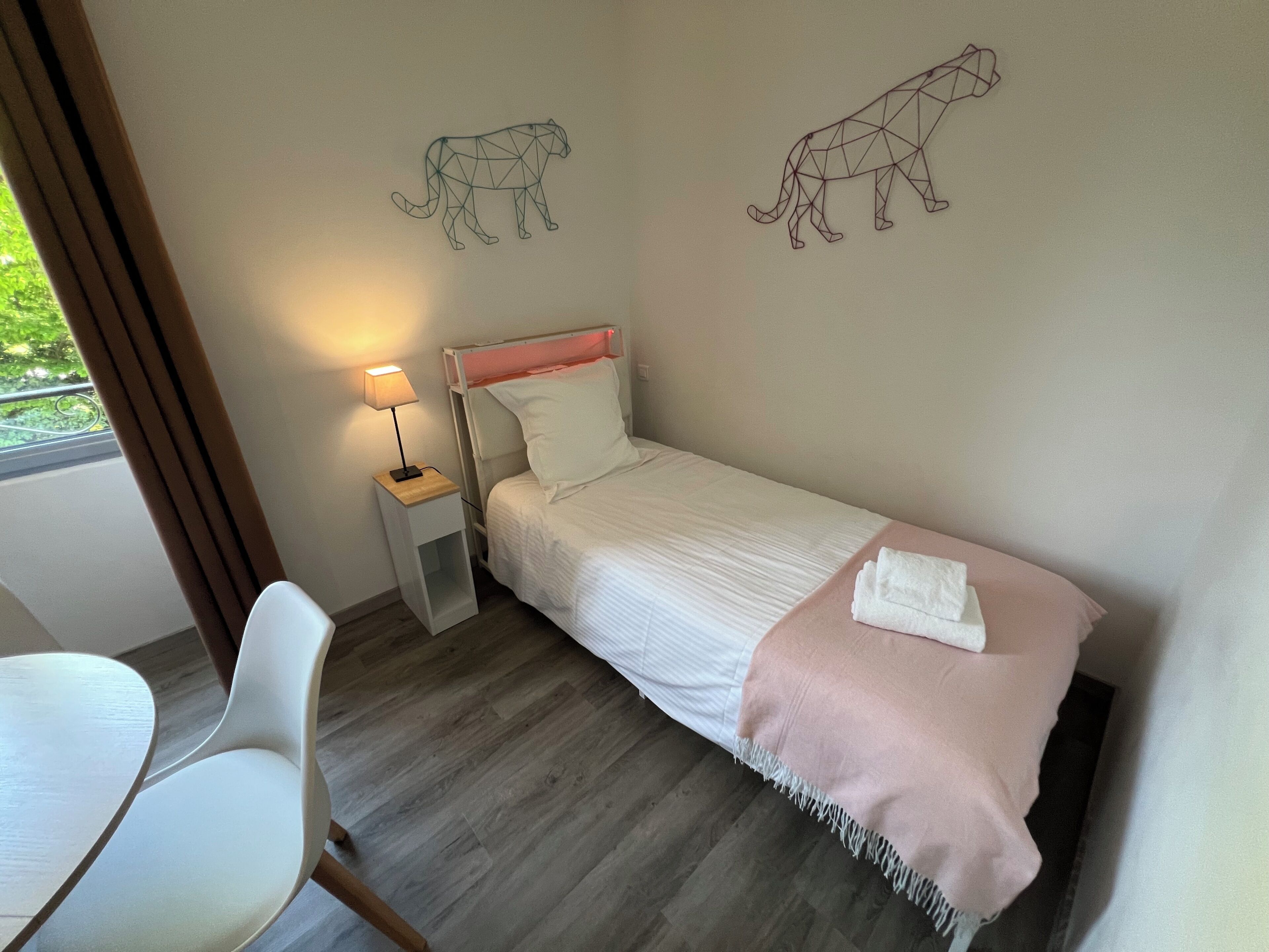 A single bed with a white and pink bedspread, a bedside table with a lamp, a chair, and two wall art pieces of a geometric animal.