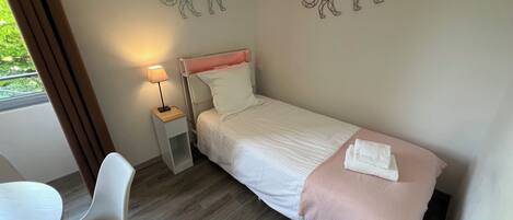 A single bed with a white and pink bedspread, a bedside table with a lamp, a chair, and two wall art pieces of a geometric animal.