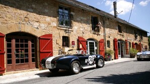 Une voiture décapotable noire d’époque est garée devant un bâtiment en pierre, doté de volets rouges et d’une enseigne indiquant qu’il s’agit d’un hôtel.