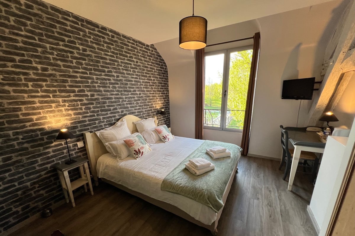 Une chambre avec un mur en briques, un lit avec des oreillers, un bureau avec une chaise et une fenêtre donnant sur un paysage agréable.