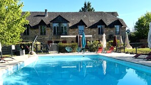 Une piscine avec des chaises longues et des parasols, un bâtiment en pierre doté de nombreuses fenêtres et un ciel dégagé.