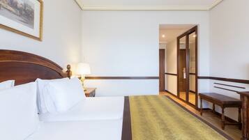 Habitació d'hotel amb un llit, una taula de nit amb una làmpada, un banc i una porta que dona a una altra habitació.